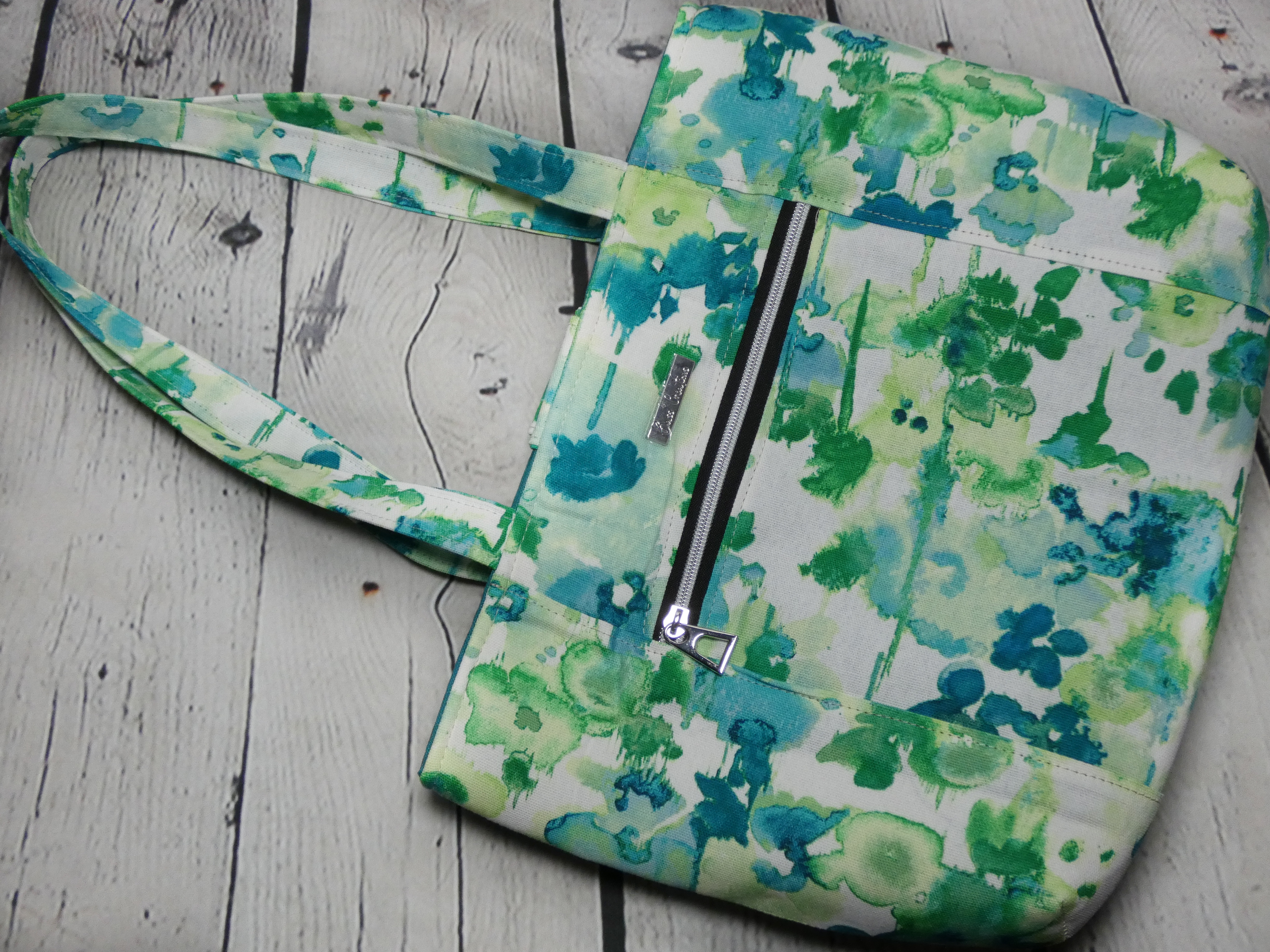 Front view of a market tote with a front zipper pocket and featuring a green splotches fabric. The zipper teeth are silver to match the silver zipper pull and silver metal Bass Creations logo.